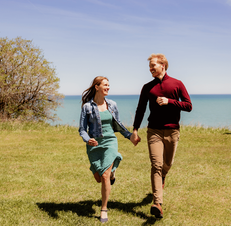 Happy couple holding hands and running through a grassy field overlooking the blue ocean on a sunny day.