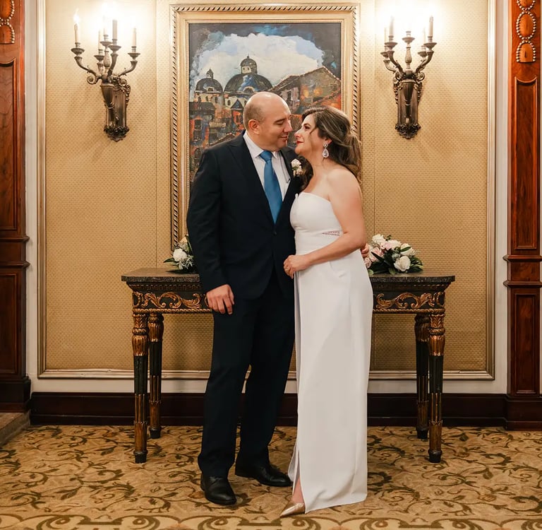 Hombre con traje junto novia con vestido blanco, viéndose mutuamente en el interior de una sala elegante con candelabros