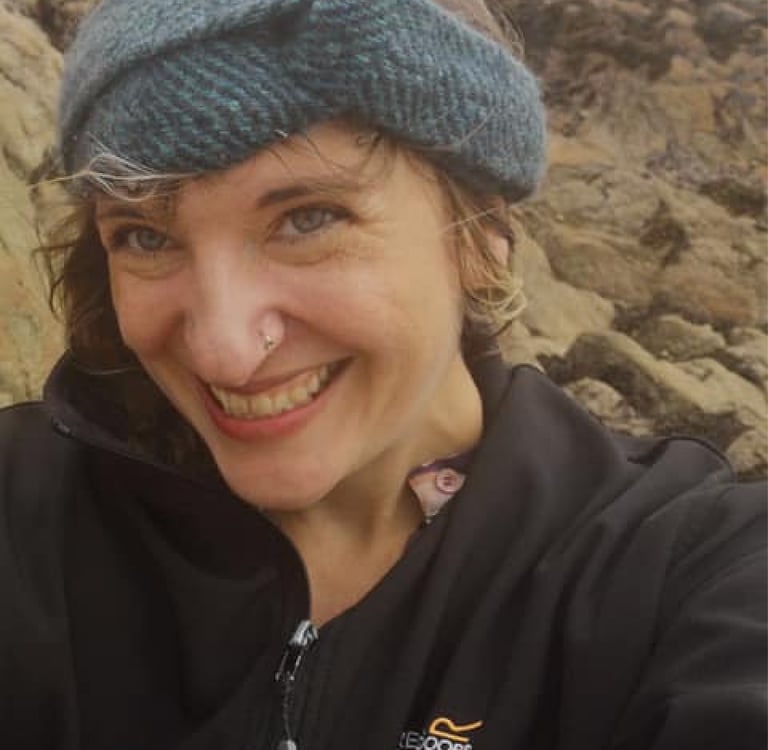 Smiling woman wearing a blue knit headband and black jacket standing on a rocky shoreline.