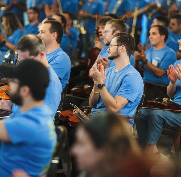 Musicians in blue t-shirts clapping during an orchestral rehearsal with violins.