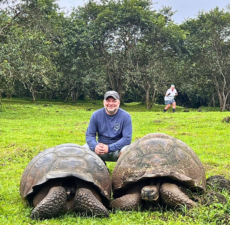 A man posing with two giant Galapagos tortoises in a lush green field near tropical trees.