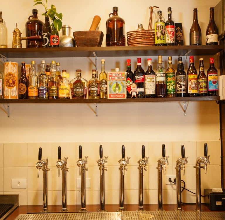 A variety of craft beer tap handles on a bar counter below shelves of liquor bottles and spirits.