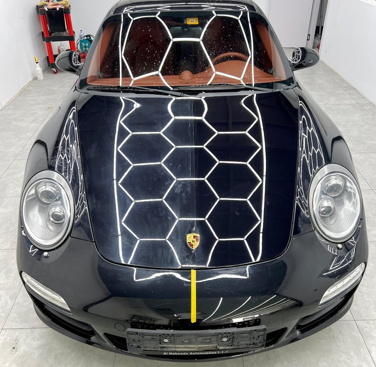 Black Porsche 911 luxury sports car with reflective honeycomb ceiling lights in a professional detailing studio.