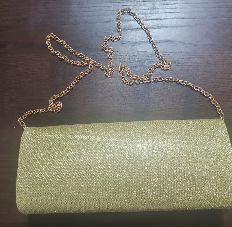 Mala clutch de festa dourada com glitter brilhante e alça de corrente metálica sobre fundo escuro.