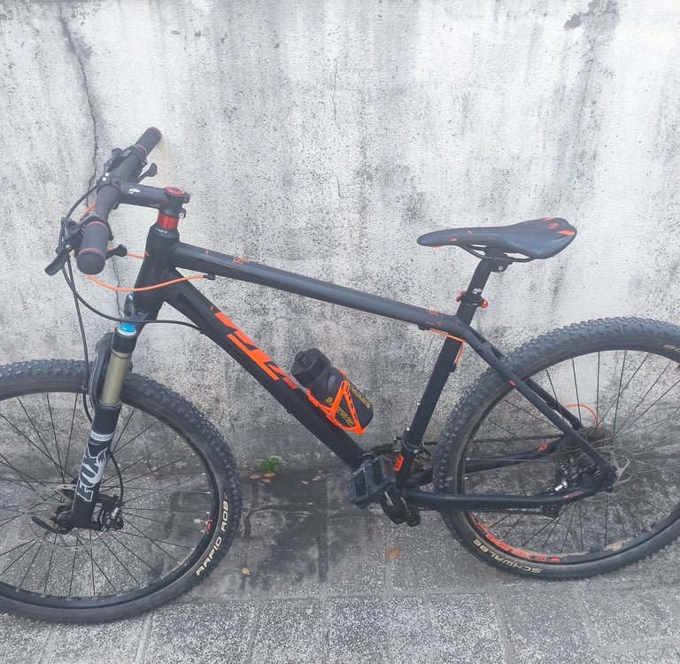 Bicicleta de montanha KTM preta com detalhes a laranja, quadro hardtail e pneus largos, vista latera