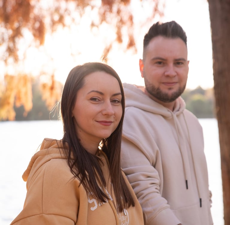 Couple wearing hoodies standing outdoors by a lake under autumn foliage at golden hour.