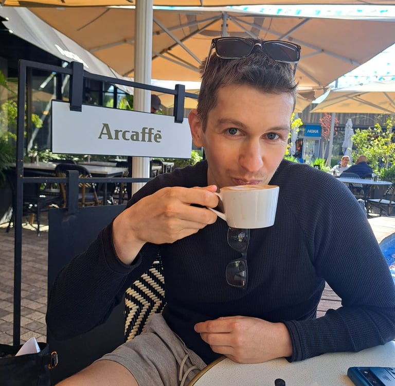 A man in a black long-sleeved shirt drinking a cappuccino at the Arcaffe outdoor cafe terrace.