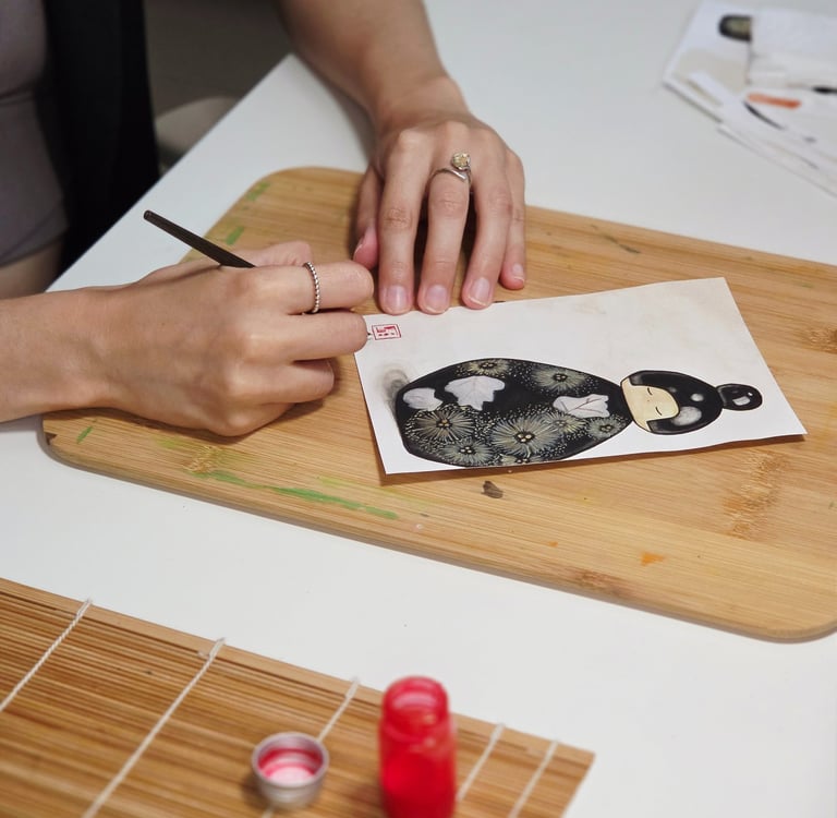 Artist painting a Japanese Kokeshi doll illustration on paper with a fine brush and red ink.