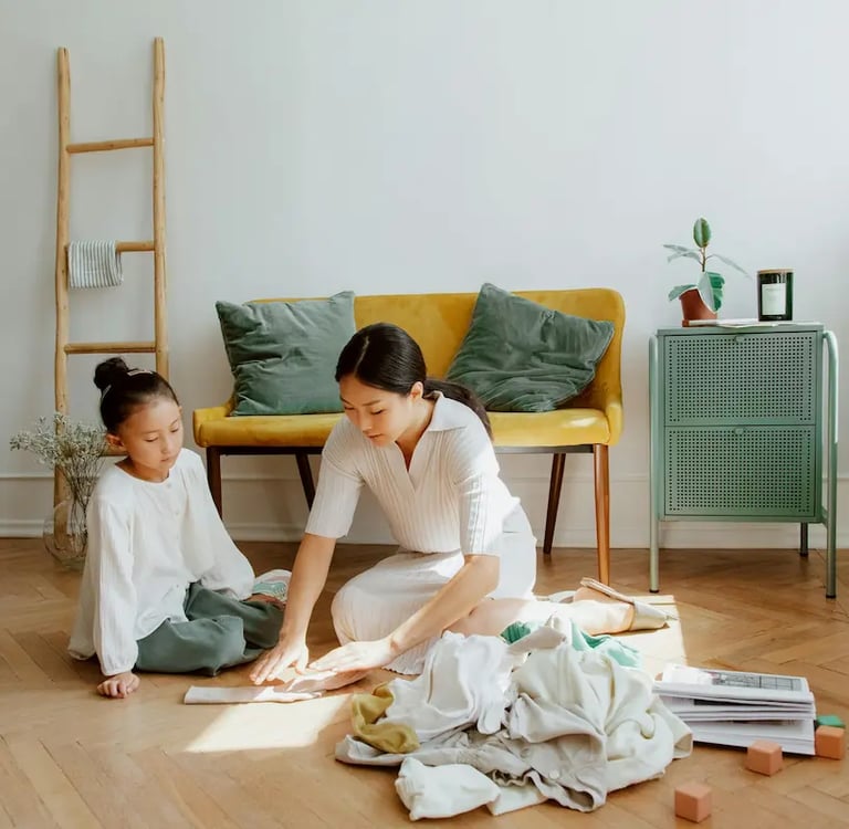 A mother and daughter organizing clothes on a wooden floor in a modern, sunlit living room.
