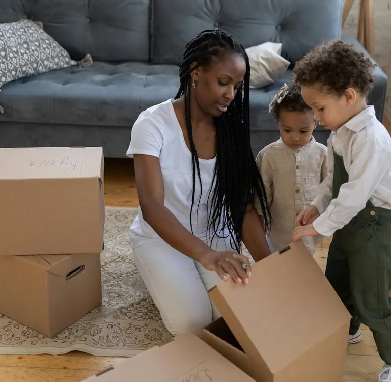 A mother and two young children unpacking cardboard boxes in their new living room.