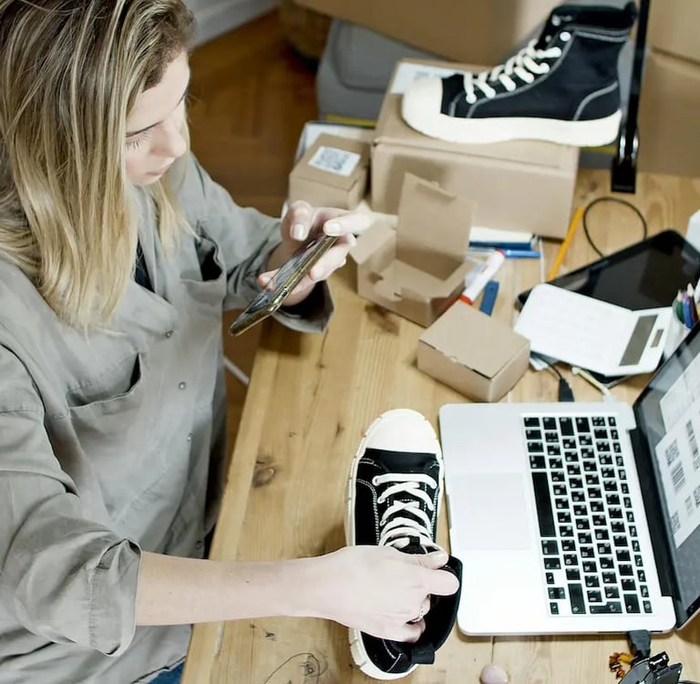 A small business owner photographs shoes on a wooden desk with a laptop and packaging, managing her online store.