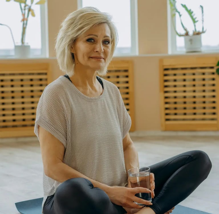A senior woman sitting on a yoga mat holding a glass of water after a workout.