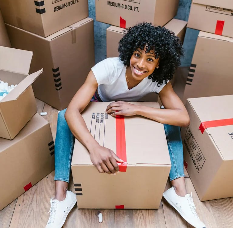 Smiling woman with curly hair sitting on the floor surrounded by cardboard moving boxes.