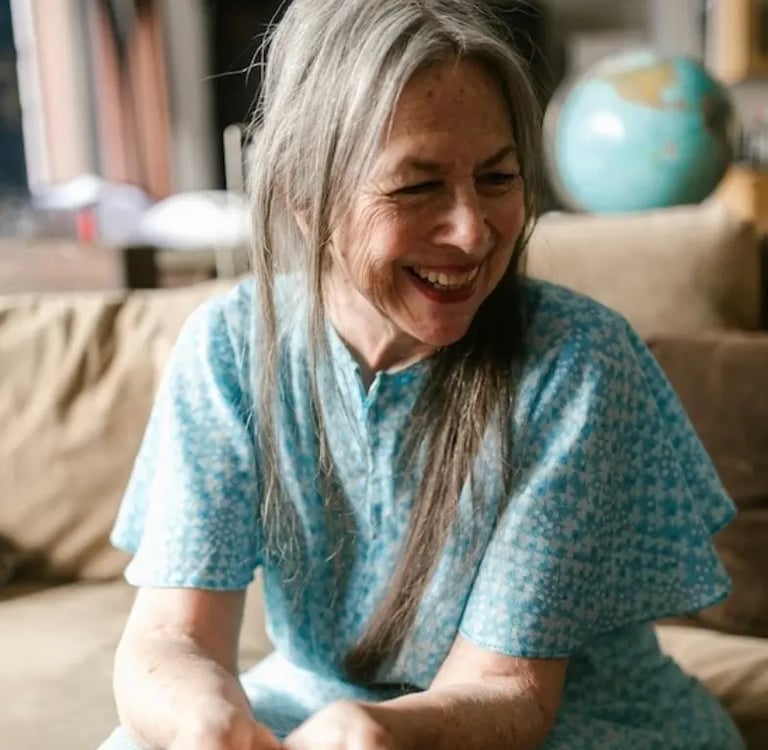 Smiling senior woman with long grey hair wearing a blue floral dress while sitting on a sofa.