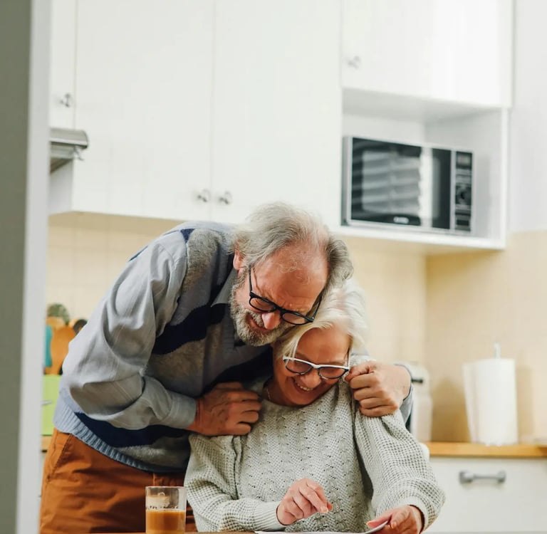 A happy senior couple at home in a bright kitchen reviewing financial documents together at a wooden table.
