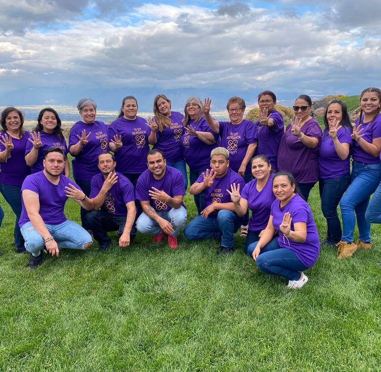 A diverse group of people wearing purple t-shirts posing on a grassy field with a mountain view.