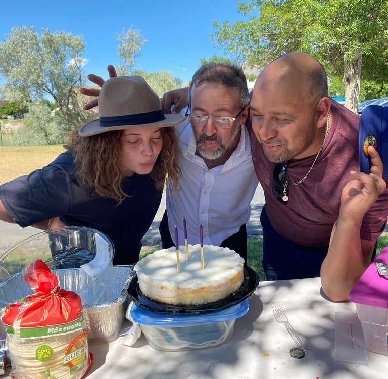 Three people blowing out purple candles on a round birthday cake at an outdoor family gathering.