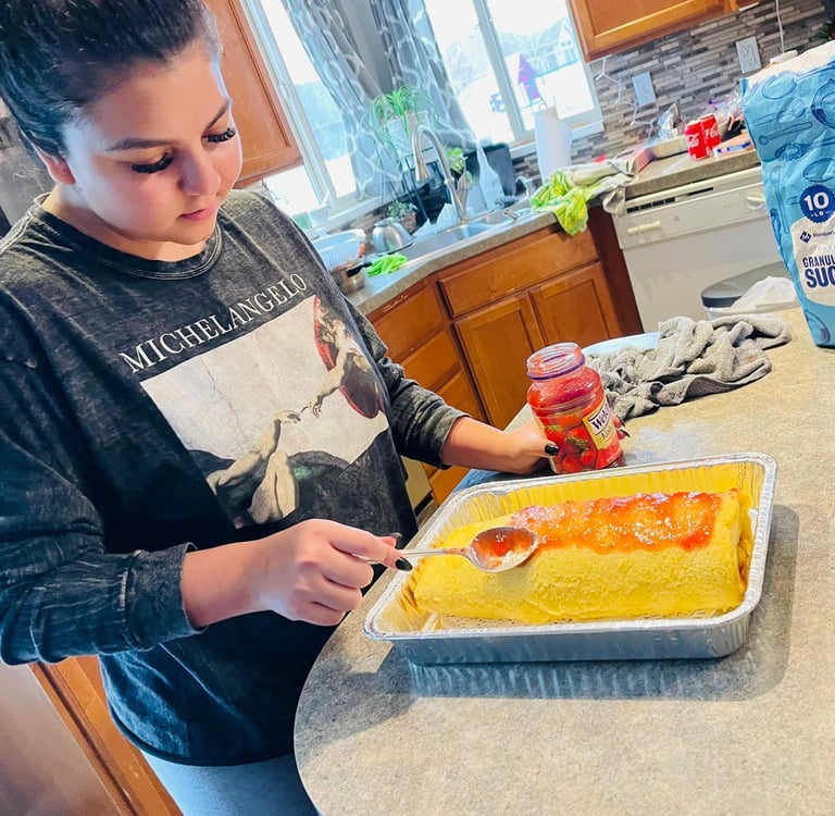 A woman spreads strawberry jam over a rolled sponge cake in a kitchen to make a Swiss roll dessert.