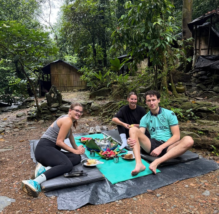 Young travelers enjoying a jungle picnic on mats near rustic wooden cabins in a tropical forest.