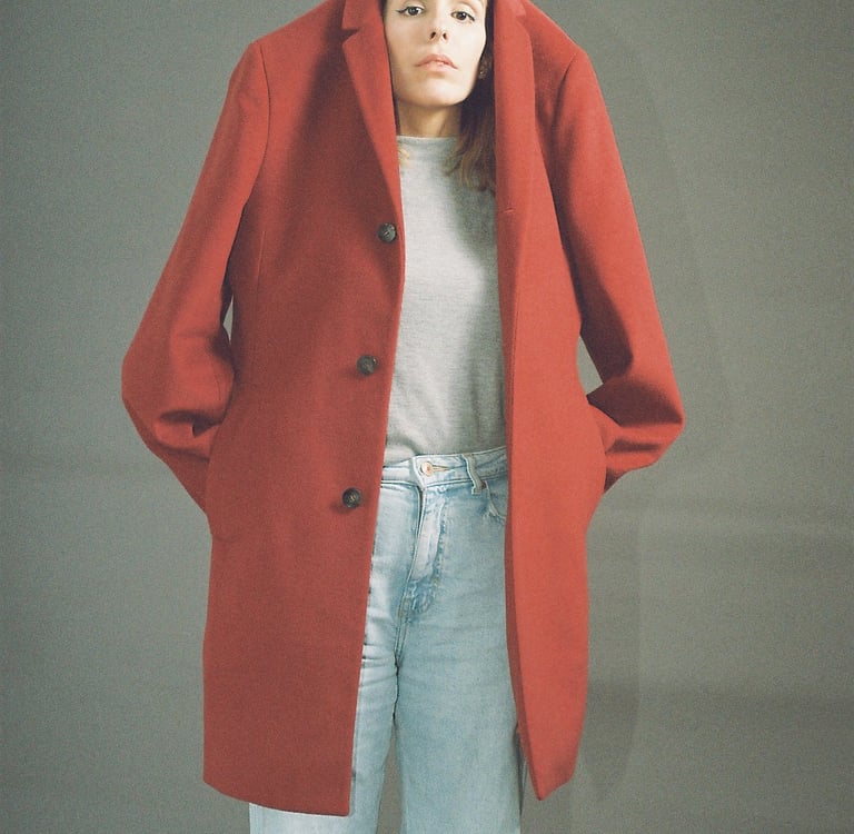 A woman wearing a large red wool coat over her head with light blue jeans against a grey background.