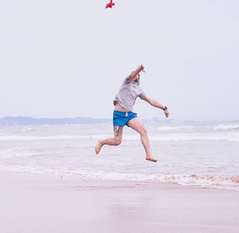A man jumps on a sandy beach while throwing a red toy glider airplane into the air.