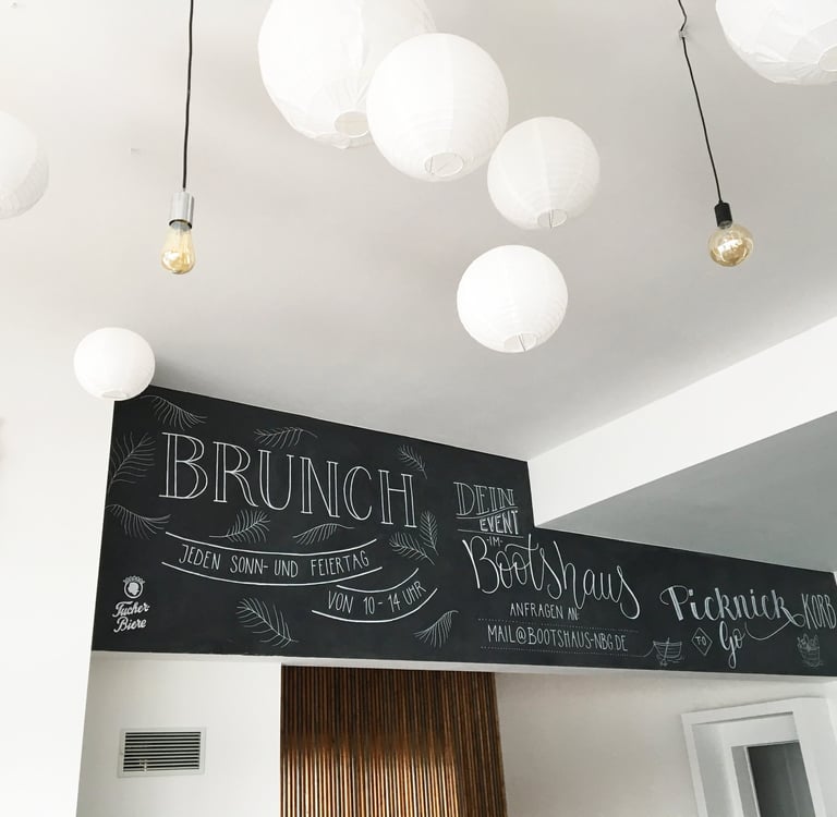 Handwritten brunch menu on a black chalkboard wall decorated with white paper lanterns and light bulbs.
