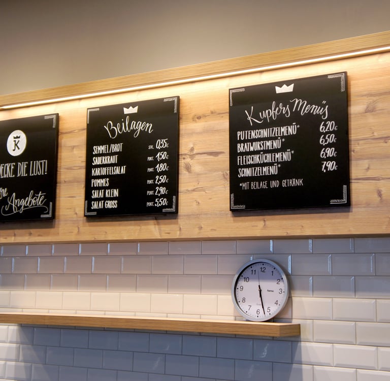German butcher shop menu boards displaying side dishes and lunch menu prices on a wooden wall above white tiles.