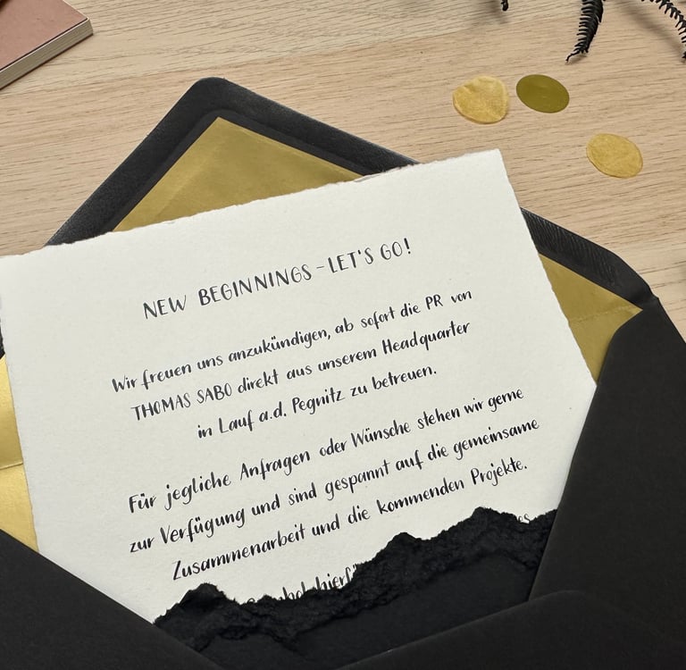 A Thomas Sabo announcement letter in a black and gold envelope on a wooden desk with a fountain pen.