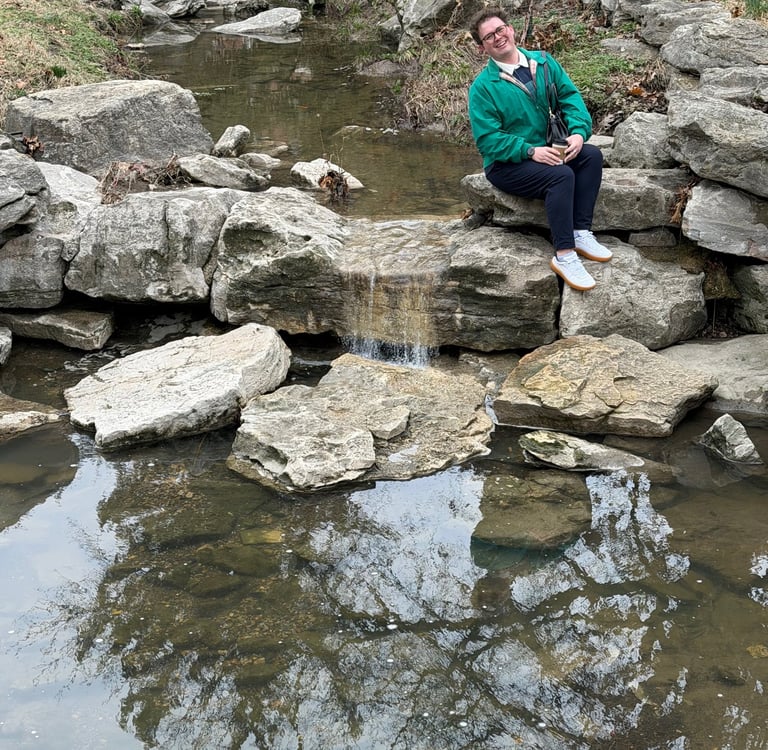 A person in a green jacket sits by a rocky stream and small waterfall in a scenic park.