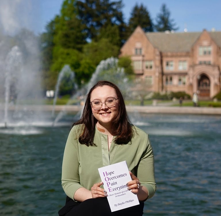 Baylie Phillips sitting in front of a fountain, posing with book Hope Overcomes Pain Everytime