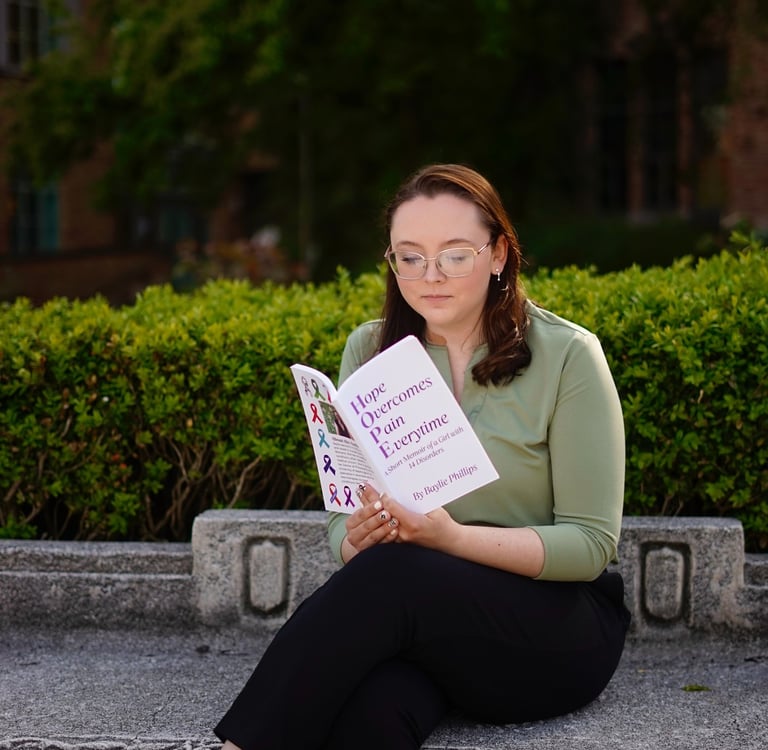 Author Baylie Phillips sitting on a bench reading Hope Overcomes Pain Overtime book.