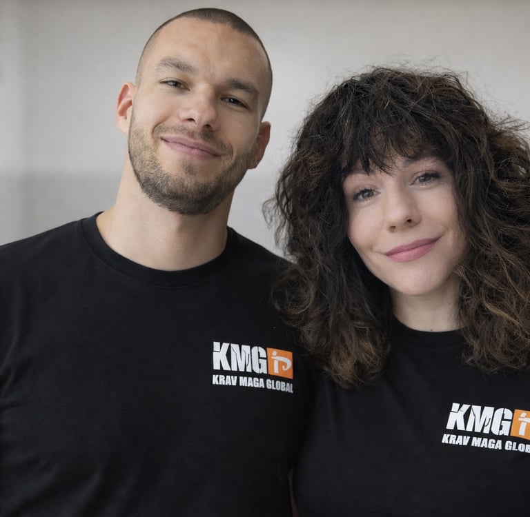 Smiling male and female Krav Maga Global instructors wearing black KMG t-shirts in a gym.