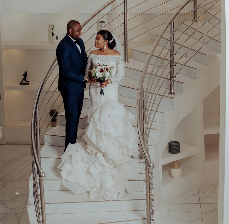 Smiling bride in a ruffled mermaid wedding dress and groom in a navy suit on a white marble spiral staircase.