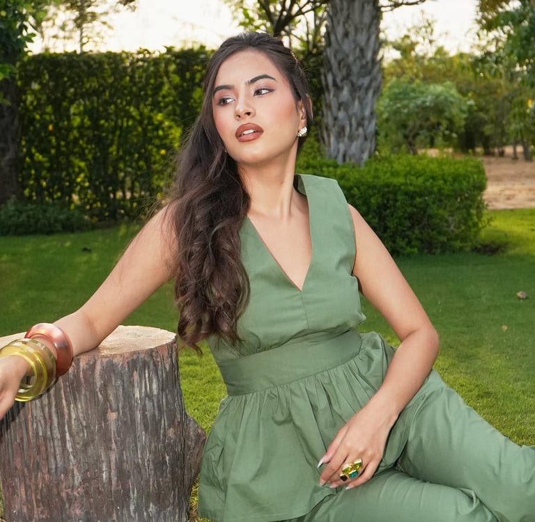 A woman in a sage green peplum jumpsuit sitting on a garden lawn leaning against a wooden tree stump.