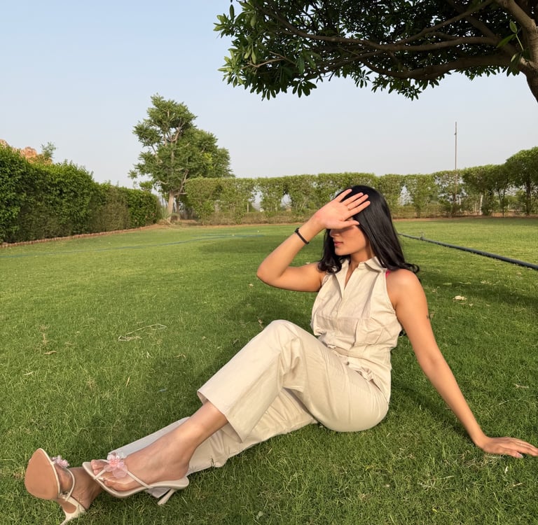 Woman in a beige jumpsuit and high heels sits on a green lawn under a tree, posing with hand over eyes.