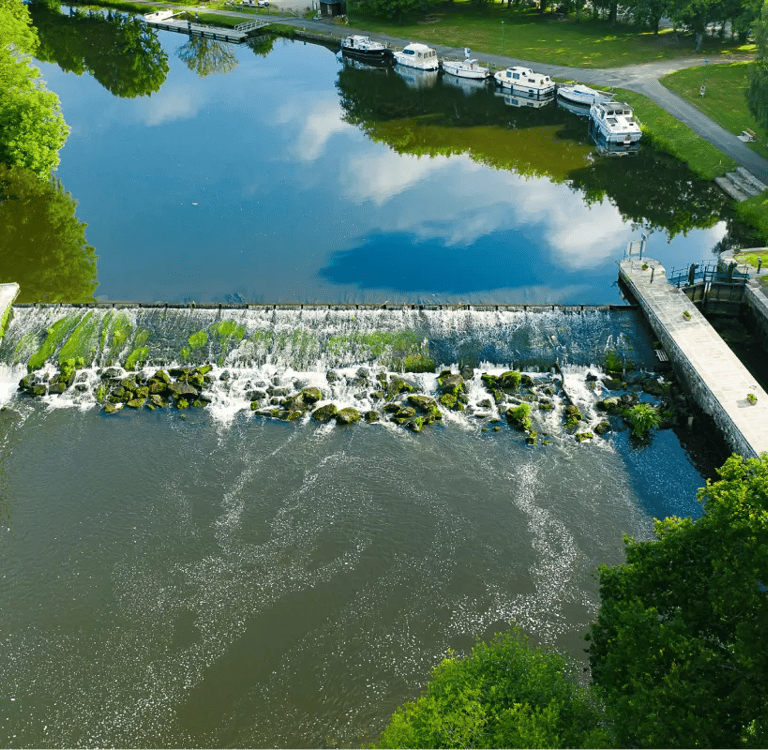 Vue aérienne d'un barrage et d'une cascade, avec des péniches amarrées et des arbres verdoyants.