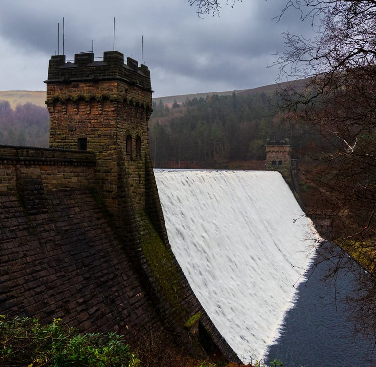 Historic stone Derwent Dam with cascading water overflow and towers in Peak District National Park.