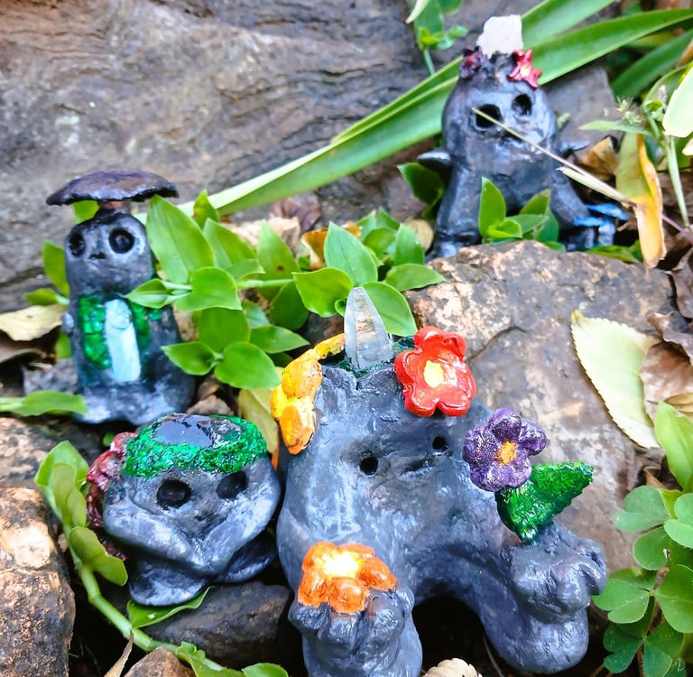 Handmade clay forest spirits with crystals and flowers sitting among garden rocks and green leaves.
