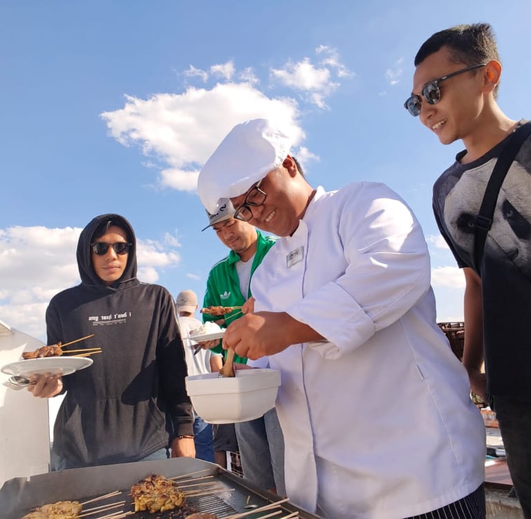 A kitchen crew grills chicken satay skewers for river crew party on a sunny outdoor deck.