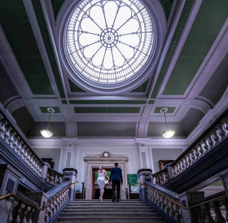 A bride and groom walk up a grand marble staircase toward a circular stained glass skylight.