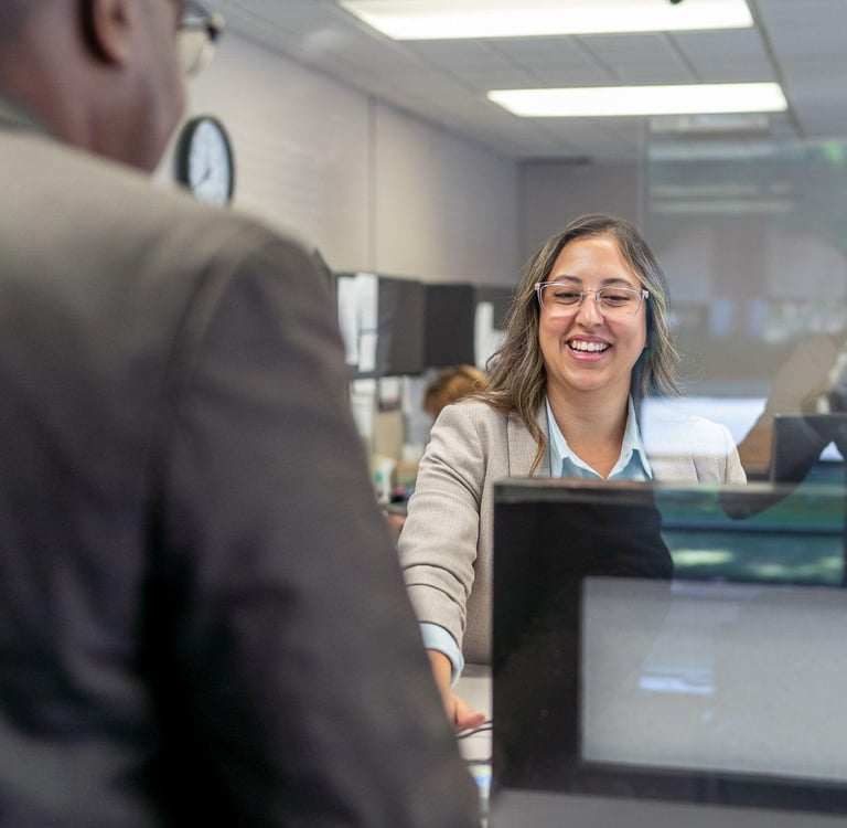 A woman in office attire smiles warmly at a customer across a reception desk.