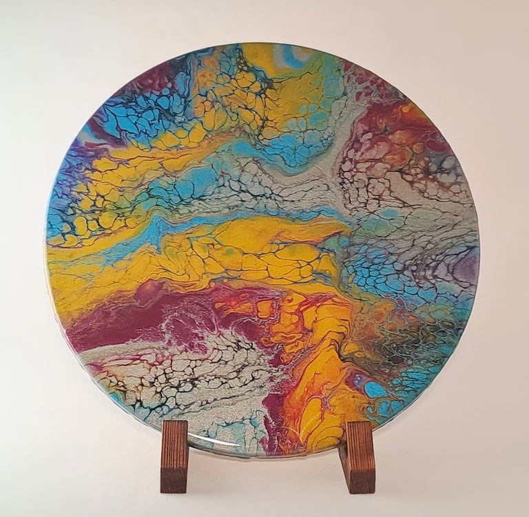 Circular acrylic pour painting with vibrant yellow, blue, and red cells on a wooden display stand.