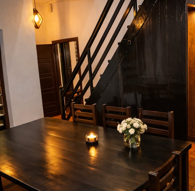 Rustic dining room with dark wood table, black staircase, candle, and white flowers in a vase.