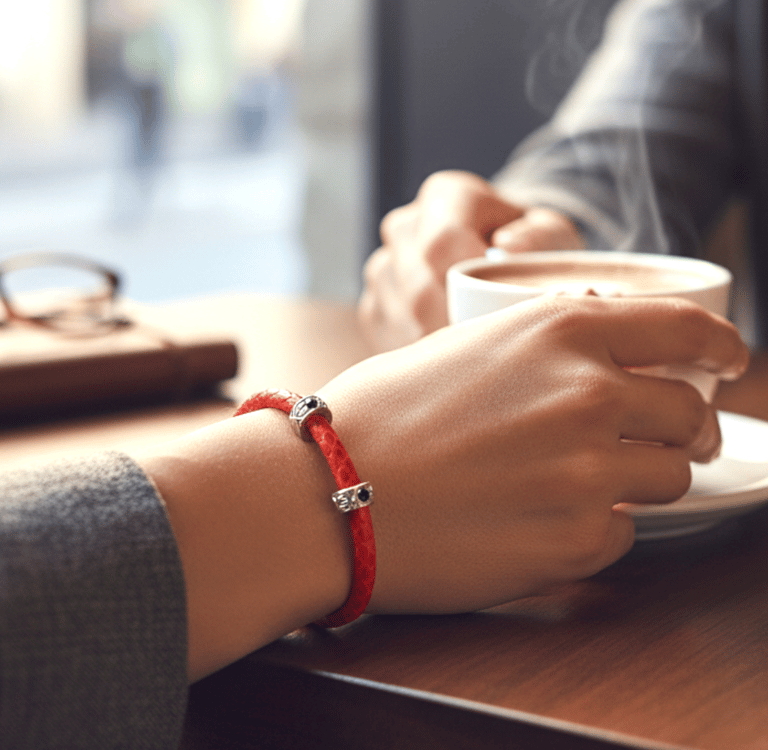 Red leather charm bracelet worn by a woman holding a steaming cup of coffee in a cafe.