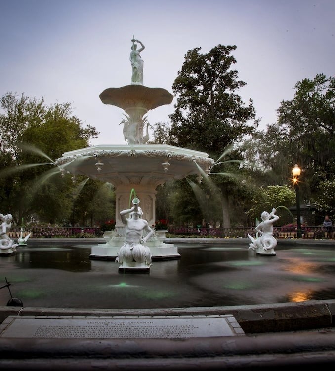 The historic Forsyth Park Fountain in Savannah, Georgia, featuring white ornate statues and spraying water.