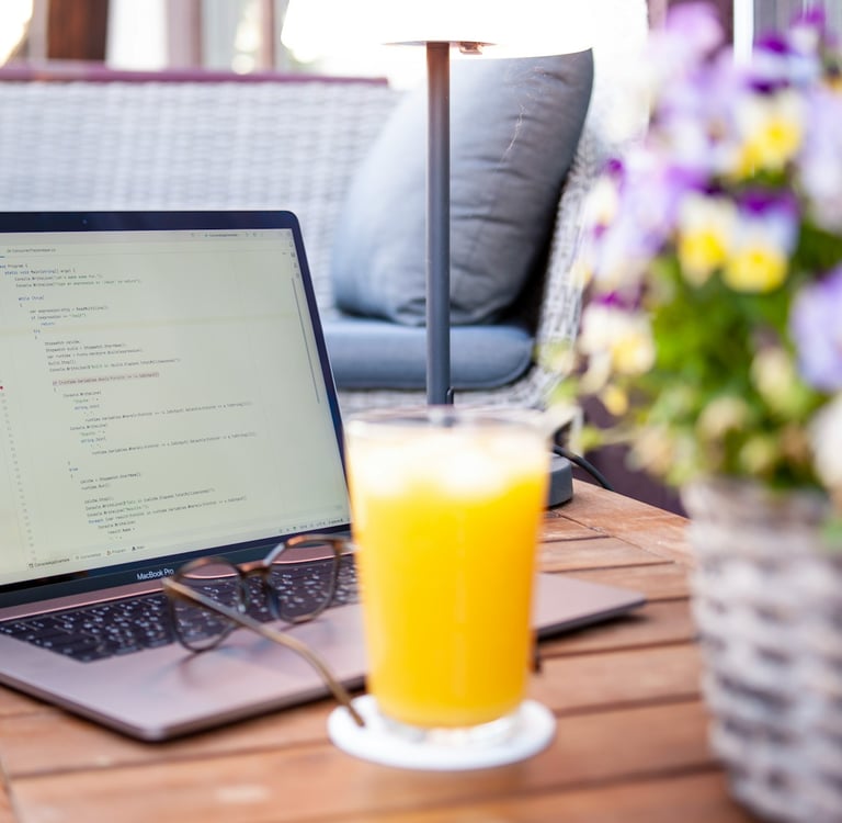 Laptop displaying computer code on a wooden outdoor table with orange juice and flowers.