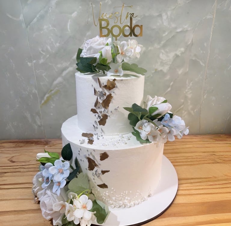 Elegante pastel de bodas blanco de dos niveles decorado con pan de oro, flores blancas y un adorno d