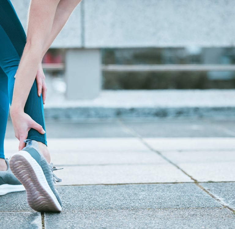 A woman in blue leggings holding her ankle due to a sports injury or sprain while running outdoors.
