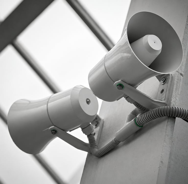 Two white public address system horn speakers mounted on a wall for announcements and audio broadcasting.