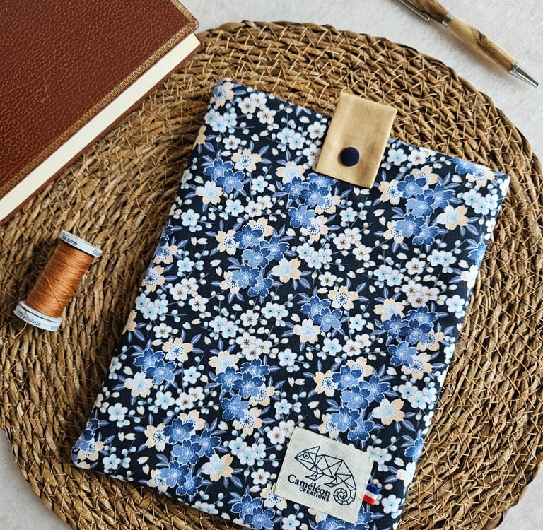 Handmade floral book sleeve with blue cherry blossom pattern, wooden pen, and leather journal on a woven mat.